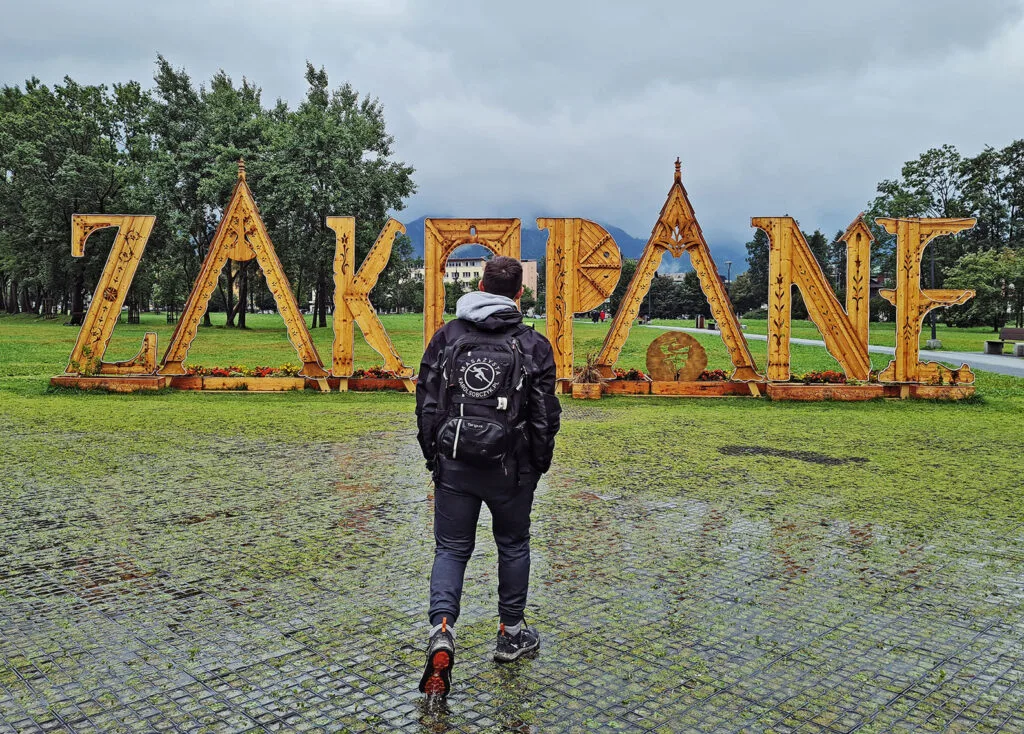 Mężczyzna z plecakiem przed drewnianym napisem Zakopane w deszczowy dzień